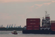 A large cargo ship is navigating the water, stacked with red shipping containers labeled 'SHIN YANG.' A smaller vessel is moving alongside by the port, leaving a trail in the water. In the background, cranes and industrial structures are visible against a somewhat cloudy sky.