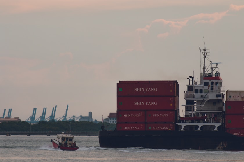 A large cargo ship is navigating the water, stacked with red shipping containers labeled 'SHIN YANG.' A smaller vessel is moving alongside by the port, leaving a trail in the water. In the background, cranes and industrial structures are visible against a somewhat cloudy sky.