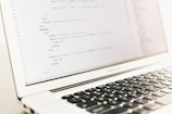 A close-up of a laptop displaying lines of HTML or programming code on its screen. The focus is on the code, with a shallow depth of field that blurs the background. The laptop is placed on a light-colored surface with its keyboard partially visible.
