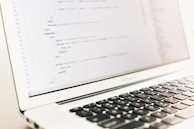 A close-up of a laptop displaying lines of HTML or programming code on its screen. The focus is on the code, with a shallow depth of field that blurs the background. The laptop is placed on a light-colored surface with its keyboard partially visible.