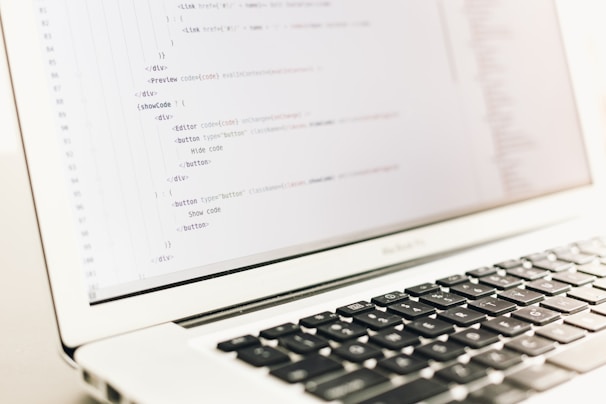 A close-up of a laptop displaying lines of HTML or programming code on its screen. The focus is on the code, with a shallow depth of field that blurs the background. The laptop is placed on a light-colored surface with its keyboard partially visible.