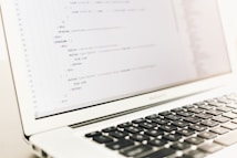 A close-up of a laptop displaying lines of HTML or programming code on its screen. The focus is on the code, with a shallow depth of field that blurs the background. The laptop is placed on a light-colored surface with its keyboard partially visible.