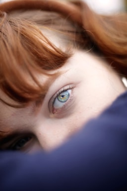 A close-up view of a person's eye with light blue and green hues, partially covered by the person's auburn hair and a dark blue material.