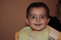 A smiling baby with wide eyes wearing a bib. The background is a neutral shade with soft lighting. The baby appears to be joyful and engaging with the camera.
