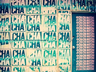 A wall covered in repeated posters with the word 'CHA' printed multiple times. The posters have a distressed, layered look and vary in shades of green and cream. To the right, a door is visible with hashtags and text '@CHA CHA MATCHA' printed in a column in a contrasting light color. The door has a chain lock.
