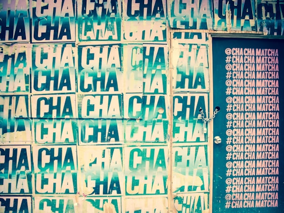 A wall covered in repeated posters with the word 'CHA' printed multiple times. The posters have a distressed, layered look and vary in shades of green and cream. To the right, a door is visible with hashtags and text '@CHA CHA MATCHA' printed in a column in a contrasting light color. The door has a chain lock.