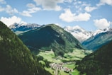 A picturesque valley surrounded by lush green hills and distant snow-capped mountains. The foreground features a small village nestled in the valley, with houses scattered among the verdant landscape. Above, a bright sky adorned with fluffy white clouds enhances the serene atmosphere.