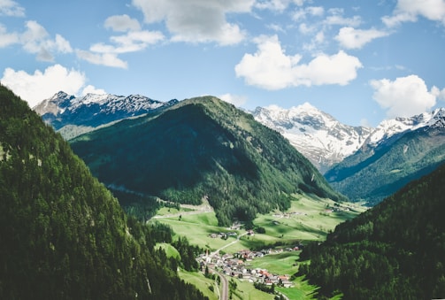 A picturesque valley surrounded by lush green hills and distant snow-capped mountains. The foreground features a small village nestled in the valley, with houses scattered among the verdant landscape. Above, a bright sky adorned with fluffy white clouds enhances the serene atmosphere.
