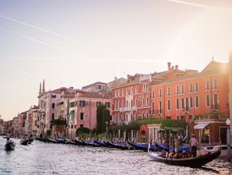 A scenic view of a canal lined with historic buildings in warm, earthy tones. Gondolas glide along the water, with people enjoying a leisurely ride. The sky is clear with subtle contrails, and the sunlight casts a soft glow over the scene.