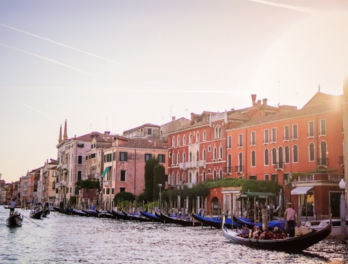 A scenic view of a canal lined with historic buildings in warm, earthy tones. Gondolas glide along the water, with people enjoying a leisurely ride. The sky is clear with subtle contrails, and the sunlight casts a soft glow over the scene.