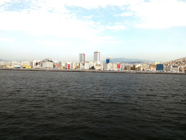 A coastal city skyline with tall buildings lined along the waterfront. The sea stretches across the foreground, and there is a mountainous landscape in the background with a partly cloudy sky above. Several buildings display large Turkish flags.