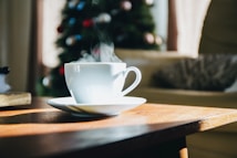 A steaming cup of coffee sits on a wooden table in a cozy room. In the background, a Christmas tree decorated with colorful ornaments is slightly out of focus. Sunlight streams through a window, casting warm shadows across the table and illuminating the cup.
