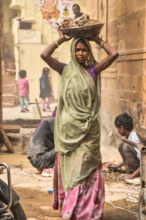 A woman wearing a green sari and pink floral skirt is carrying a large bowl of rocks on her head. She appears focused and determined. In the background, several people are engaged in laborious activities, including a person squatting near the ground and two children in colorful clothing. The area appears dusty, suggesting construction or road work, with an unfinished or rustic environment. The wall behind is made of stone and features a colorful poster with religious imagery.