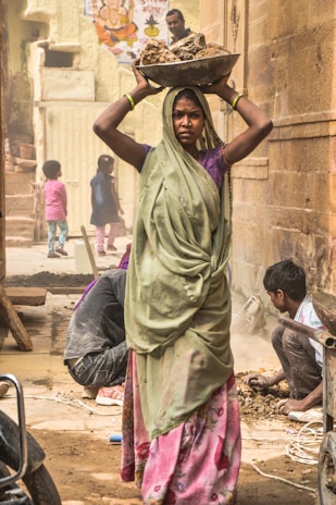 A woman wearing a green sari and pink floral skirt is carrying a large bowl of rocks on her head. She appears focused and determined. In the background, several people are engaged in laborious activities, including a person squatting near the ground and two children in colorful clothing. The area appears dusty, suggesting construction or road work, with an unfinished or rustic environment. The wall behind is made of stone and features a colorful poster with religious imagery.