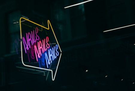 A brightly lit neon sign with the word 'NAILS' repeated multiple times in vibrant colors including pink, blue, and white. The sign is arranged in an arrow shape pointing downward, set against a dark background, likely a window or glass surface reflecting some soft lights.