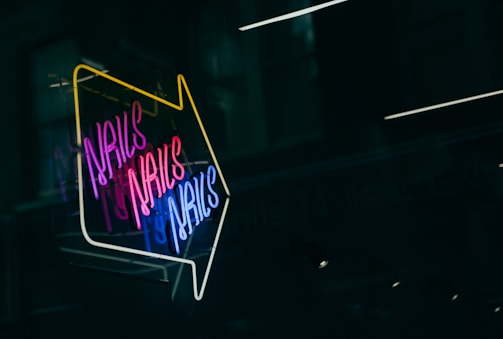 A brightly lit neon sign with the word 'NAILS' repeated multiple times in vibrant colors including pink, blue, and white. The sign is arranged in an arrow shape pointing downward, set against a dark background, likely a window or glass surface reflecting some soft lights.