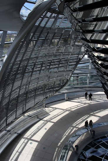 An architectural interior with curved glass and metal structures casting intricate shadows on the floor. The space is open and airy, with a few people standing and sitting on a circular platform.