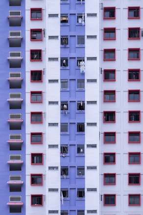 A high-rise residential building facade featuring a pattern of red-framed windows against a backdrop of purple and white walls. Windows are open with visible laundry hanging out to dry, suggesting a dense urban living environment.