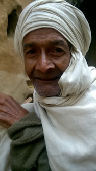 An older man with a gentle smile, wearing a white turban and robe, is captured in the image. The background has earthy tones, as if taken in a rustic or rural setting.