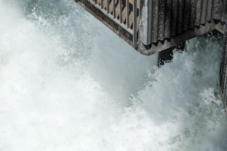 A powerful cascade of water splashes against a reinforced metal structure, which appears to be part of a bridge or walkway. The water is turbulent and foamy, creating a dynamic and energetic scene.