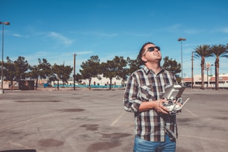 A person wearing sunglasses and a checkered shirt is holding a drone controller and looking upwards, standing in a parking lot with clear blue skies. Trees and a few tall streetlights are visible in the background, along with some commercial buildings and signs.