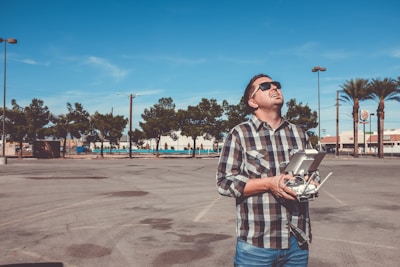 A person wearing sunglasses and a checkered shirt is holding a drone controller and looking upwards, standing in a parking lot with clear blue skies. Trees and a few tall streetlights are visible in the background, along with some commercial buildings and signs.