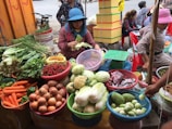 A busy market stall features a variety of fresh produce displayed in colorful baskets. Vegetables such as carrots, onions, cabbage, eggplants, chili peppers, and leafy greens are neatly arranged. Two women are attending to the produce, with one woman peeling corn while wearing a blue hat.