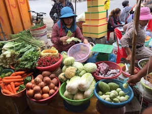 A busy market stall features a variety of fresh produce displayed in colorful baskets. Vegetables such as carrots, onions, cabbage, eggplants, chili peppers, and leafy greens are neatly arranged. Two women are attending to the produce, with one woman peeling corn while wearing a blue hat.