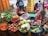 A busy market stall features a variety of fresh produce displayed in colorful baskets. Vegetables such as carrots, onions, cabbage, eggplants, chili peppers, and leafy greens are neatly arranged. Two women are attending to the produce, with one woman peeling corn while wearing a blue hat.