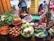 A busy market stall features a variety of fresh produce displayed in colorful baskets. Vegetables such as carrots, onions, cabbage, eggplants, chili peppers, and leafy greens are neatly arranged. Two women are attending to the produce, with one woman peeling corn while wearing a blue hat.
