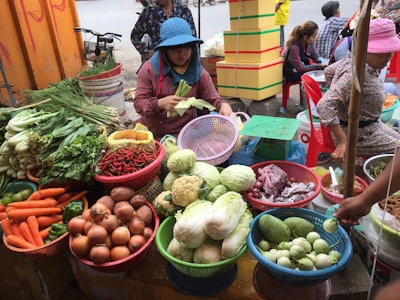 A busy market stall features a variety of fresh produce displayed in colorful baskets. Vegetables such as carrots, onions, cabbage, eggplants, chili peppers, and leafy greens are neatly arranged. Two women are attending to the produce, with one woman peeling corn while wearing a blue hat.