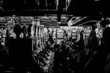 A dimly lit casino filled with rows of slot machines, some occupied by people. The machines' lights contrast with the dark surroundings, creating a vibrant yet subdued atmosphere.