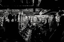 A dimly lit casino filled with rows of slot machines, some occupied by people. The machines' lights contrast with the dark surroundings, creating a vibrant yet subdued atmosphere.