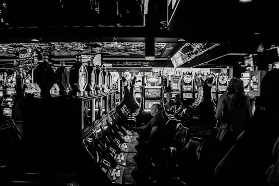 A dimly lit casino filled with rows of slot machines, some occupied by people. The machines' lights contrast with the dark surroundings, creating a vibrant yet subdued atmosphere.