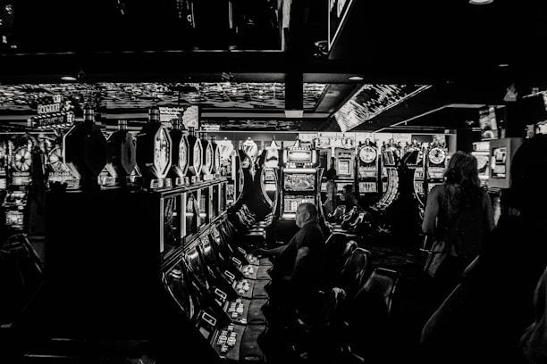 A dimly lit casino filled with rows of slot machines, some occupied by people. The machines' lights contrast with the dark surroundings, creating a vibrant yet subdued atmosphere.