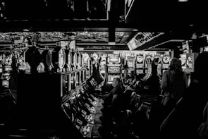 A dimly lit casino filled with rows of slot machines, some occupied by people. The machines' lights contrast with the dark surroundings, creating a vibrant yet subdued atmosphere.
