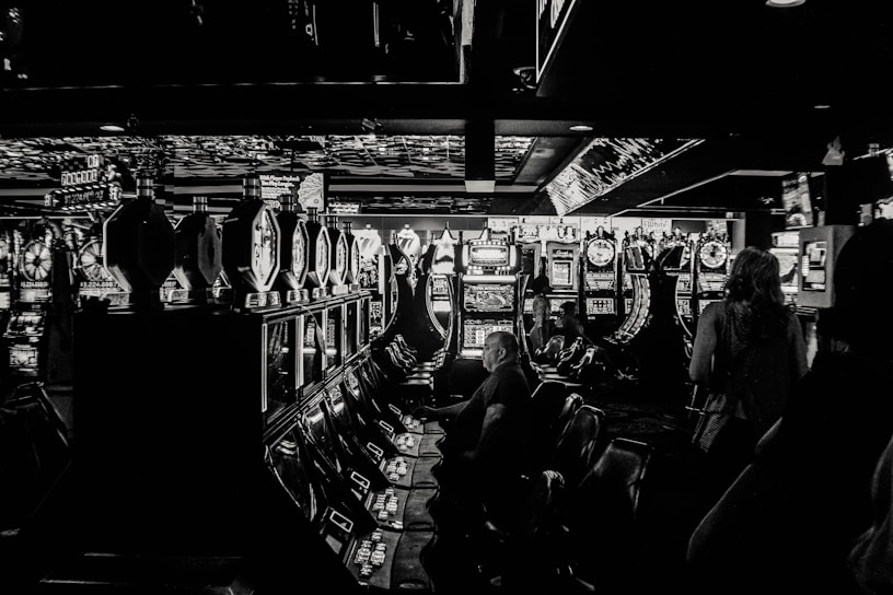 A dimly lit casino filled with rows of slot machines, some occupied by people. The machines' lights contrast with the dark surroundings, creating a vibrant yet subdued atmosphere.