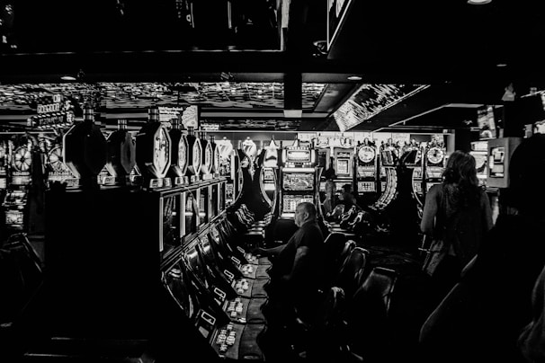 A dimly lit casino filled with rows of slot machines, some occupied by people. The machines' lights contrast with the dark surroundings, creating a vibrant yet subdued atmosphere.