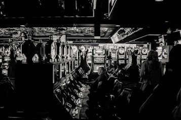 A dimly lit casino filled with rows of slot machines, some occupied by people. The machines' lights contrast with the dark surroundings, creating a vibrant yet subdued atmosphere.