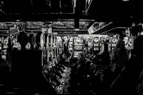 A dimly lit casino filled with rows of slot machines, some occupied by people. The machines' lights contrast with the dark surroundings, creating a vibrant yet subdued atmosphere.