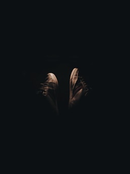 A pair of sneakers illuminated in dim lighting, creating a stark contrast against the dark background. The focus is on the shoes, highlighting their texture and design.