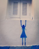 A minimalist wall mural depicting a blue silhouette of a girl in a polka dot dress reaching up toward a closed white shuttered window. The painting is simple and uses bold blue hues against a textured off-white wall, creating a striking contrast.