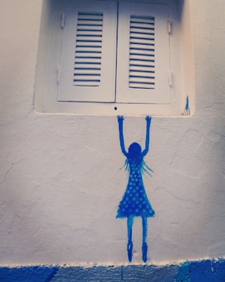 A minimalist wall mural depicting a blue silhouette of a girl in a polka dot dress reaching up toward a closed white shuttered window. The painting is simple and uses bold blue hues against a textured off-white wall, creating a striking contrast.