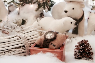 A wooden watch rests on a burlap pillow inside a wooden box, surrounded by snow-covered decorations, including a pine cone, white twigs tied with rope, and plush polar bear figures. The setting appears festive and wintry, suggesting a cozy holiday theme.
