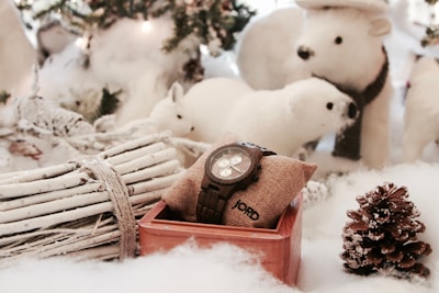 A wooden watch rests on a burlap pillow inside a wooden box, surrounded by snow-covered decorations, including a pine cone, white twigs tied with rope, and plush polar bear figures. The setting appears festive and wintry, suggesting a cozy holiday theme.