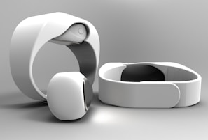 A set of sleek, modern white electronic wristbands featuring a minimalist design. The bands have a smooth, curved surface and appear to be made of a glossy material. They are positioned on a light gray background, with one band standing upright and another laid flat.