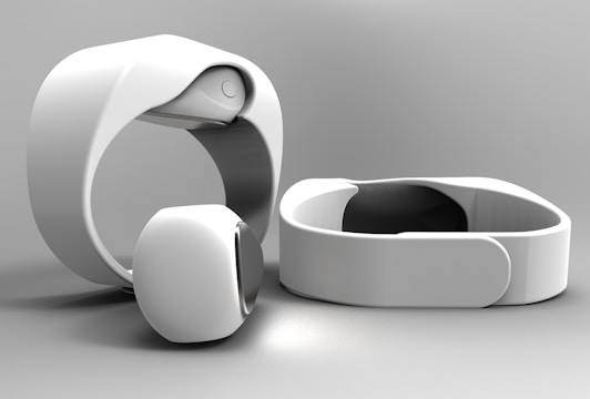 A set of sleek, modern white electronic wristbands featuring a minimalist design. The bands have a smooth, curved surface and appear to be made of a glossy material. They are positioned on a light gray background, with one band standing upright and another laid flat.