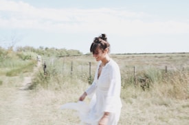A woman wearing a light, flowing white dress is walking leisurely in a grassy field under a bright sky. She appears relaxed and is smiling, with her hair tied in a loose bun. The background features a rustic fence and a pathway that extends through the landscape, conveying a serene and open environment.