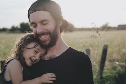 A bearded man wearing a cap embraces a young child, who is smiling and leaning against his chest. They are standing in a sunlit field with a soft focus background, creating a serene and joyful atmosphere.