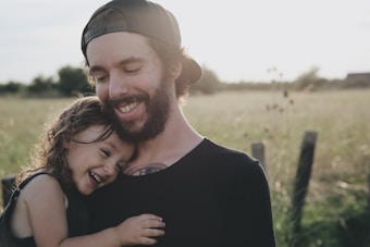 A bearded man wearing a cap embraces a young child, who is smiling and leaning against his chest. They are standing in a sunlit field with a soft focus background, creating a serene and joyful atmosphere.